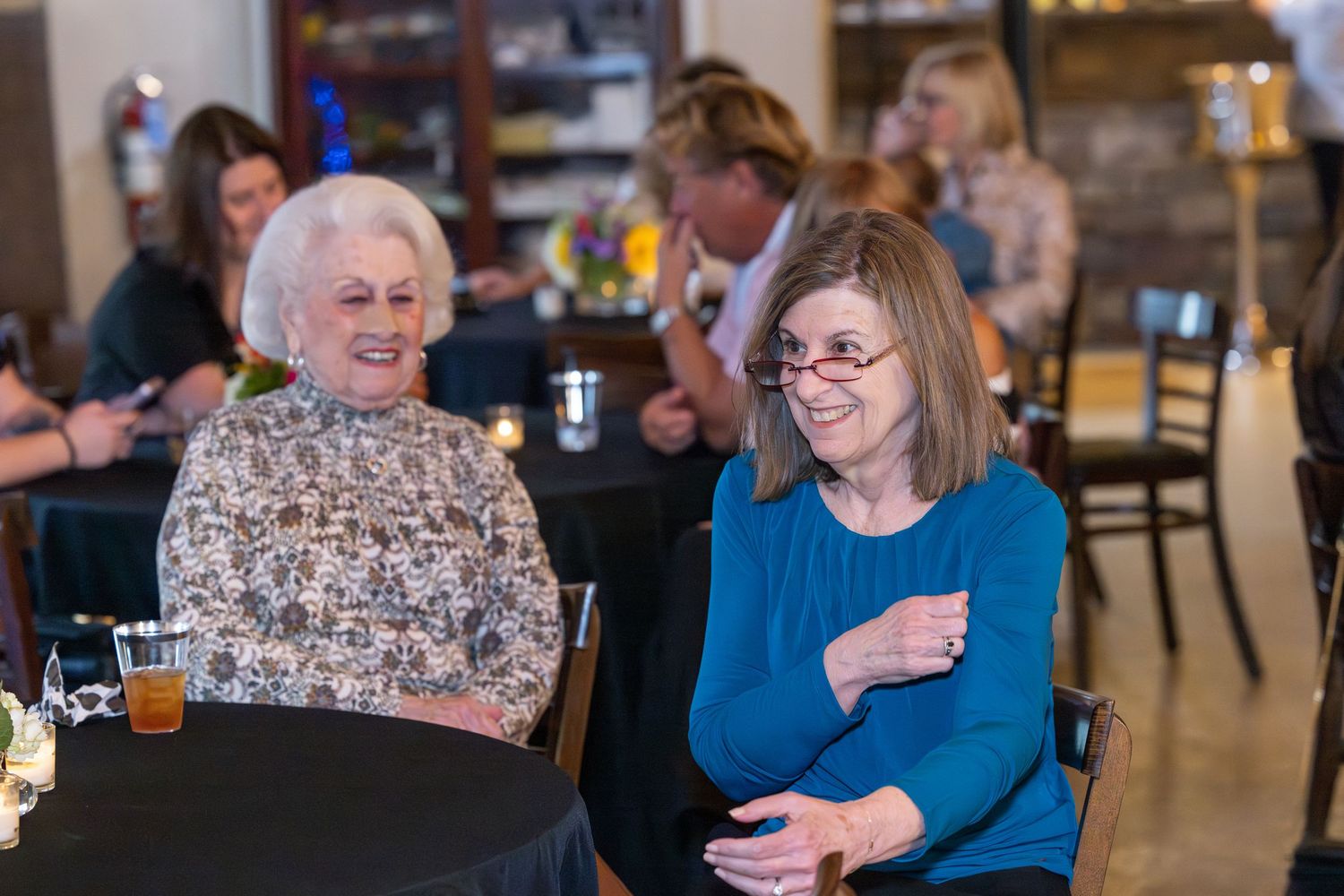 Two older women smiling and laughing together at a social gathering in a restaurant setting.