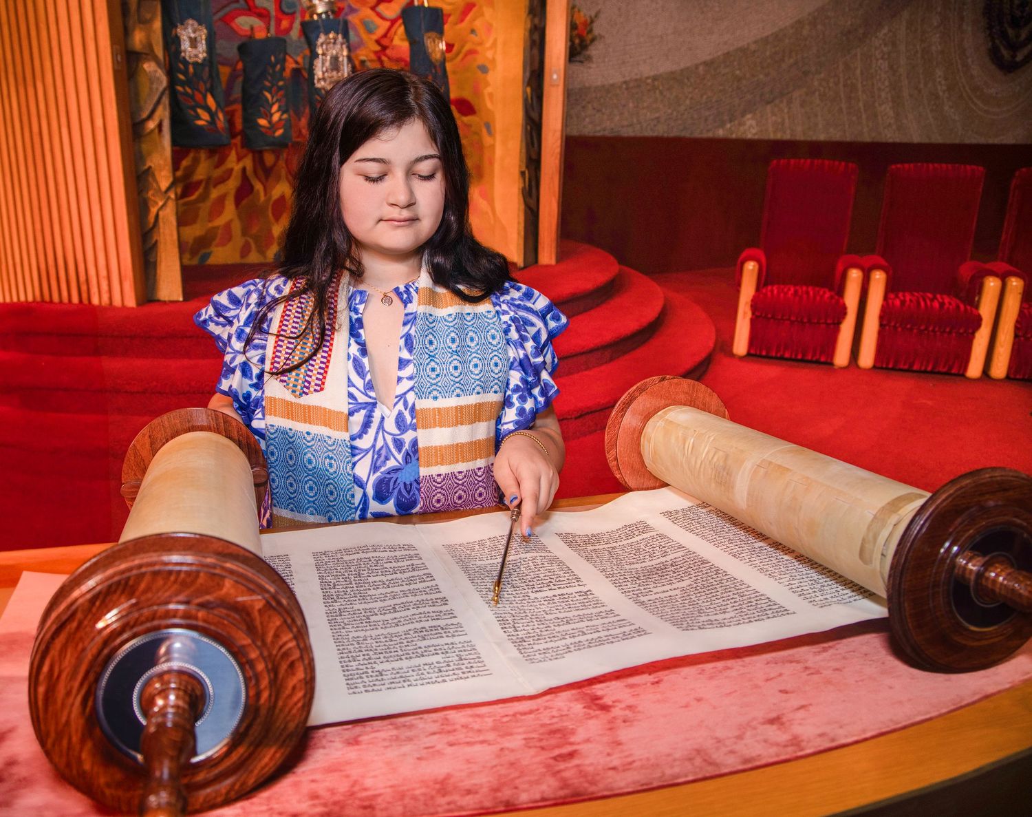 A person reads from a Torah scroll during a religious ceremony at a synagogue with red carpeting.
