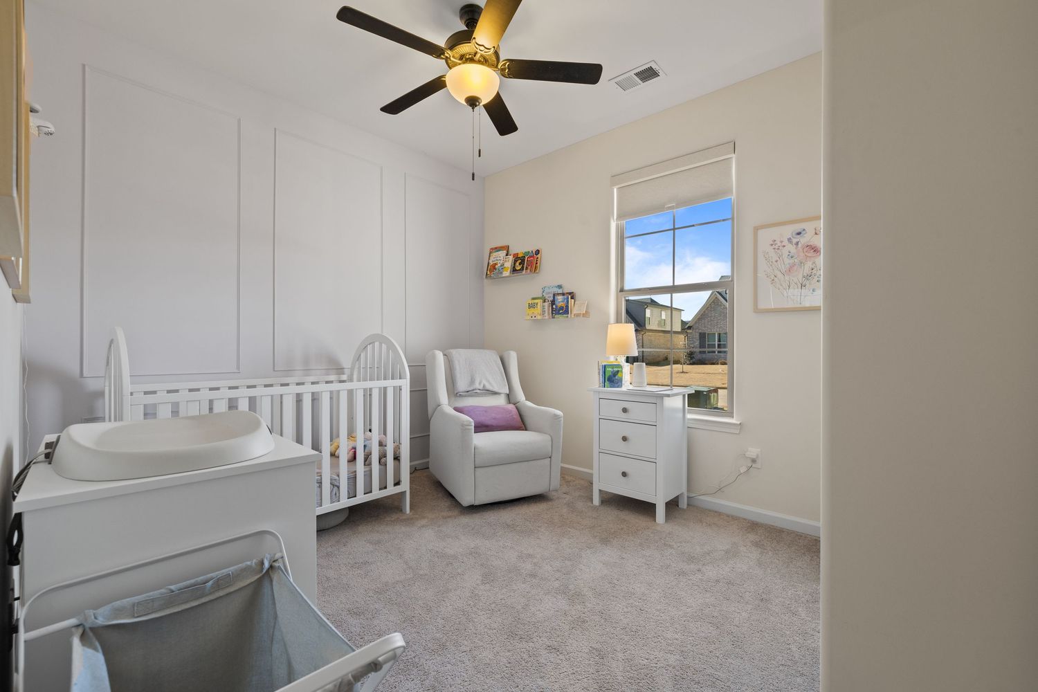 Modern nursery room with white crib, ceiling fan, and natural lighting through window features neutral decor and carpeting.