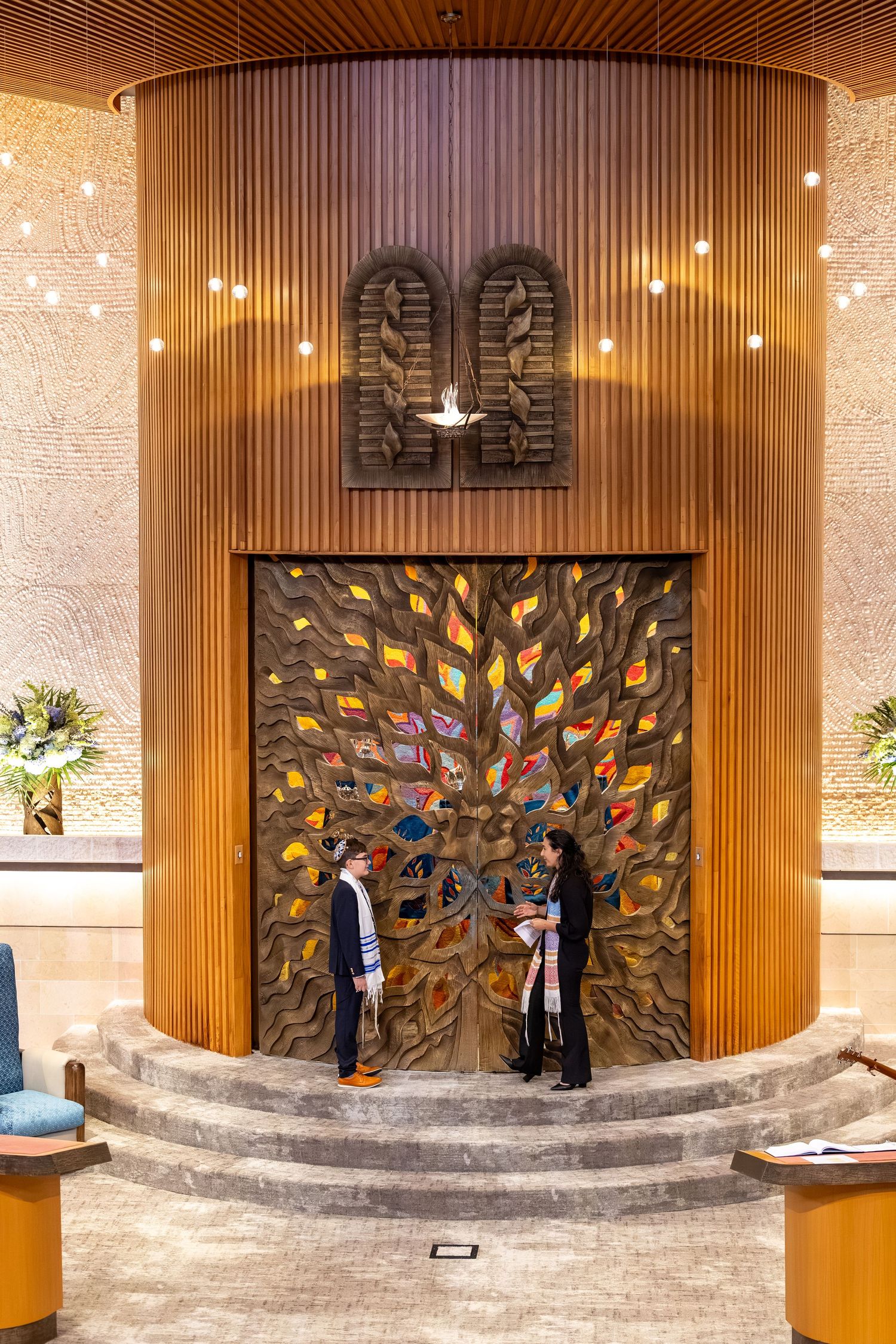Two people stand before an ornate wooden ark with colorful tree sculpture and Ten Commandments in a synagogue.