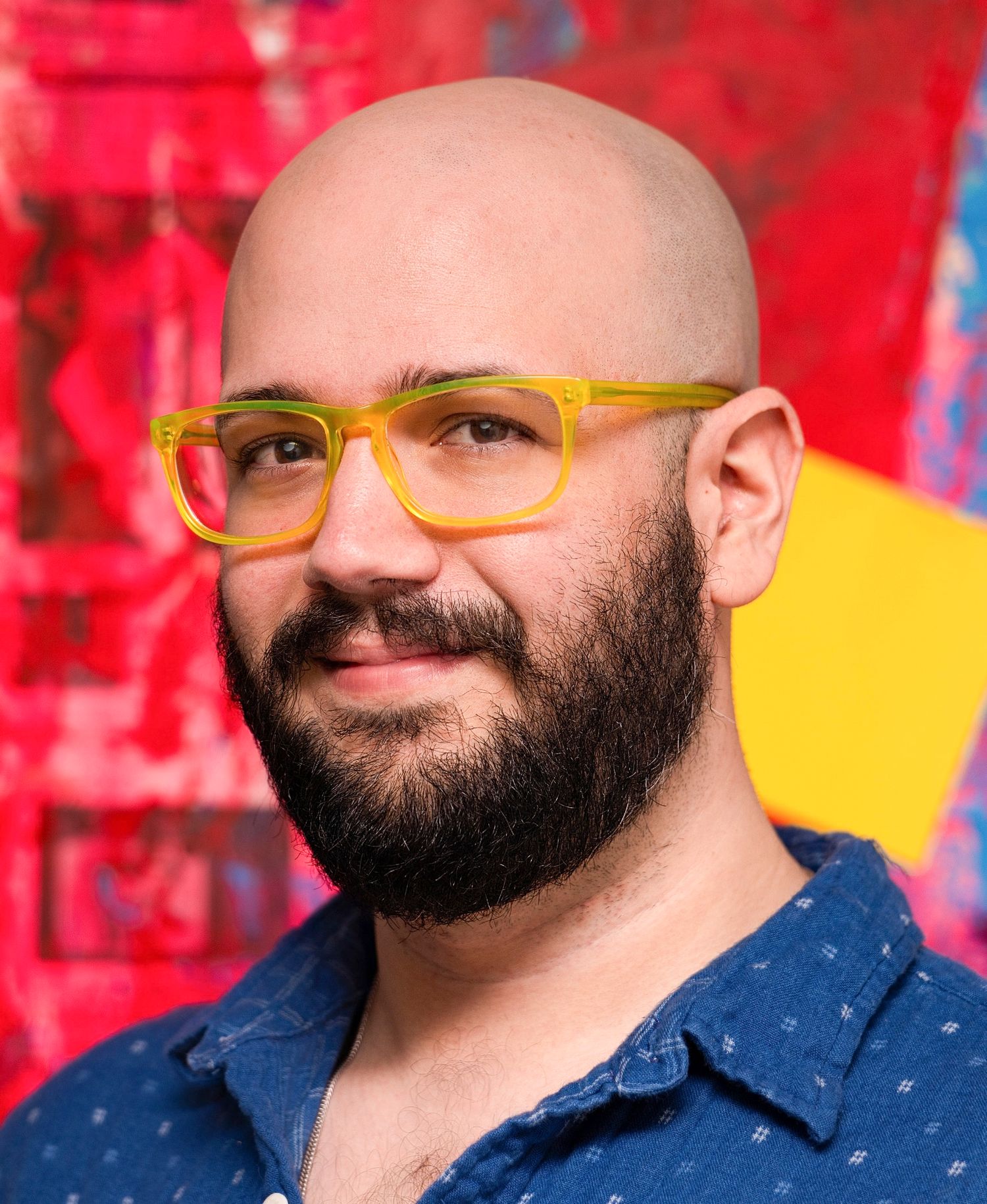 Portrait of person with beard wearing yellow glasses against colorful red background.