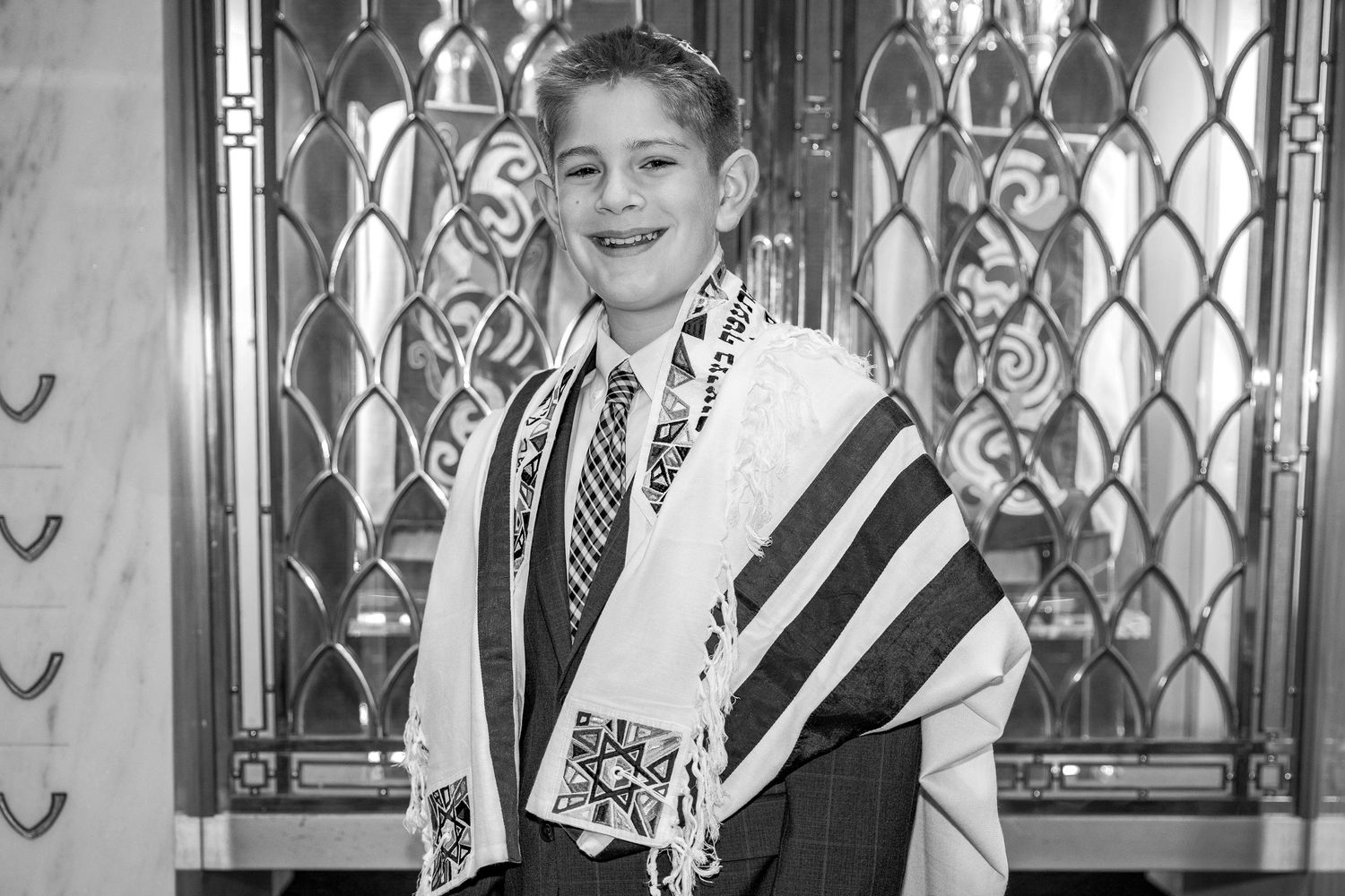 Young boy in suit and tallit smiles during his Bar Mitzvah ceremony in a synagogue.