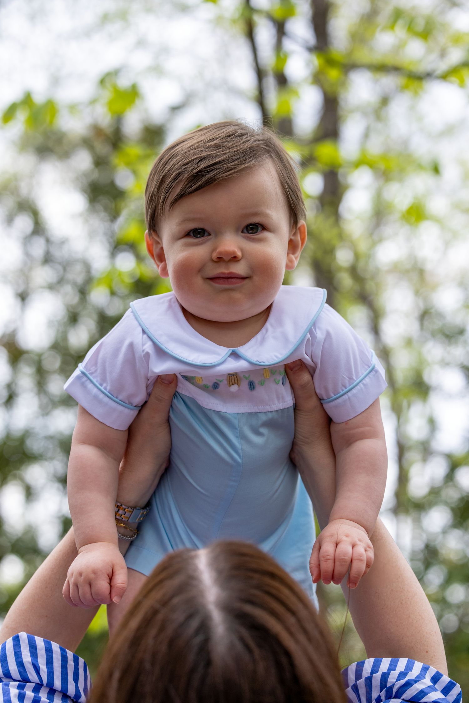 A baby wearing a light blue and white outfit smiles while being lifted up outdoors against a leafy background.