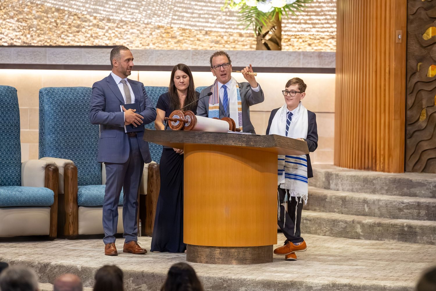 Family celebrating a Bar Mitzvah ceremony at a synagogue, with boy in tallit at podium.