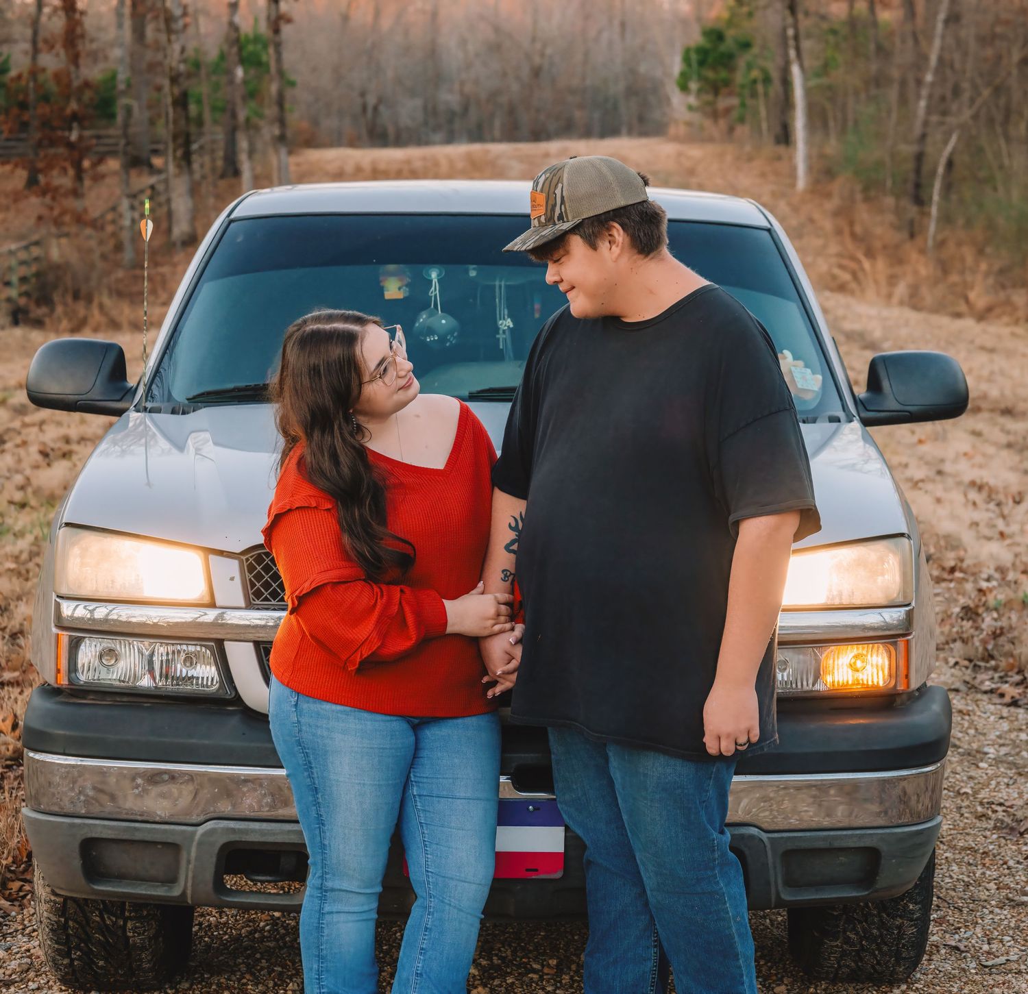 A couple stands close together in front of a parked car during sunset in a wooded area.