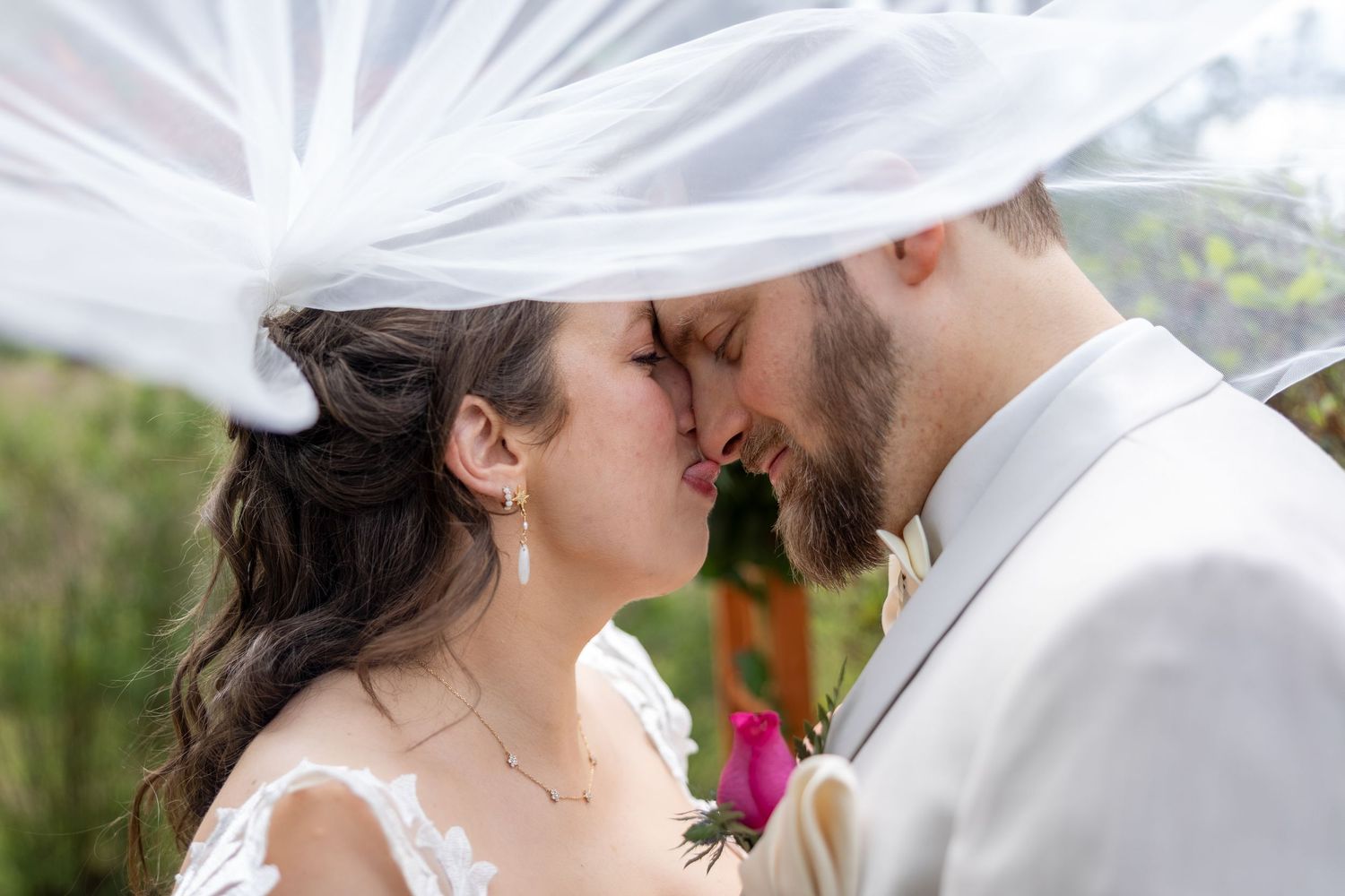 Bride and groom sharing a romantic kiss under a flowing white veil outdoors on their wedding day.