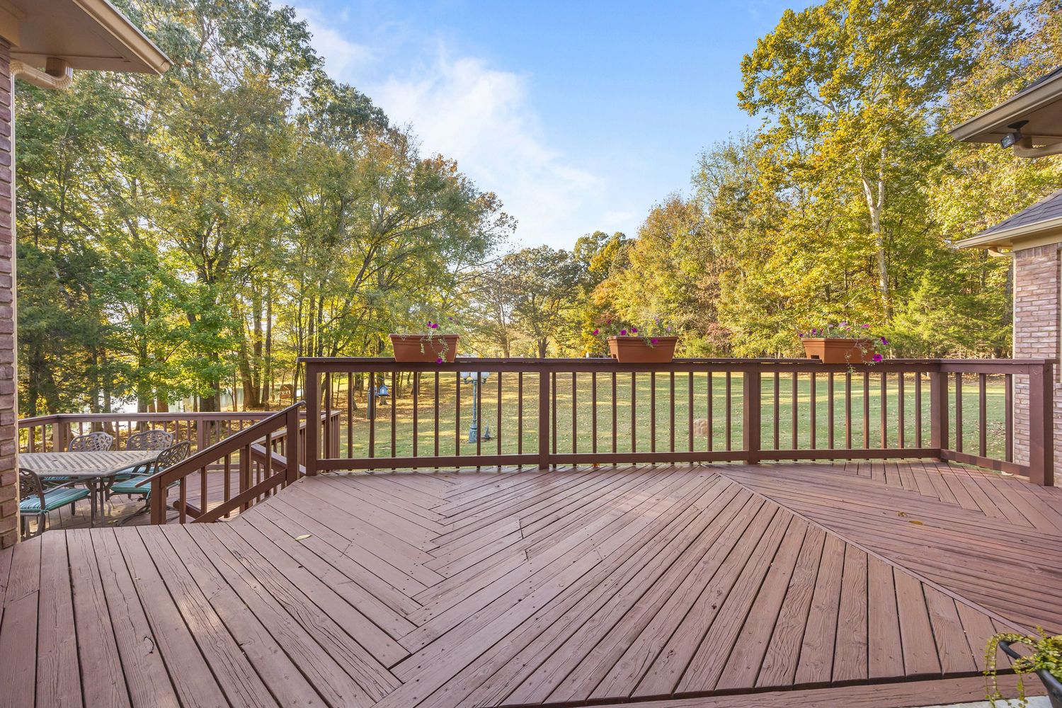 Large wooden deck with dark brown staining and black railings overlooking a scenic wooded backyard on a sunny day.