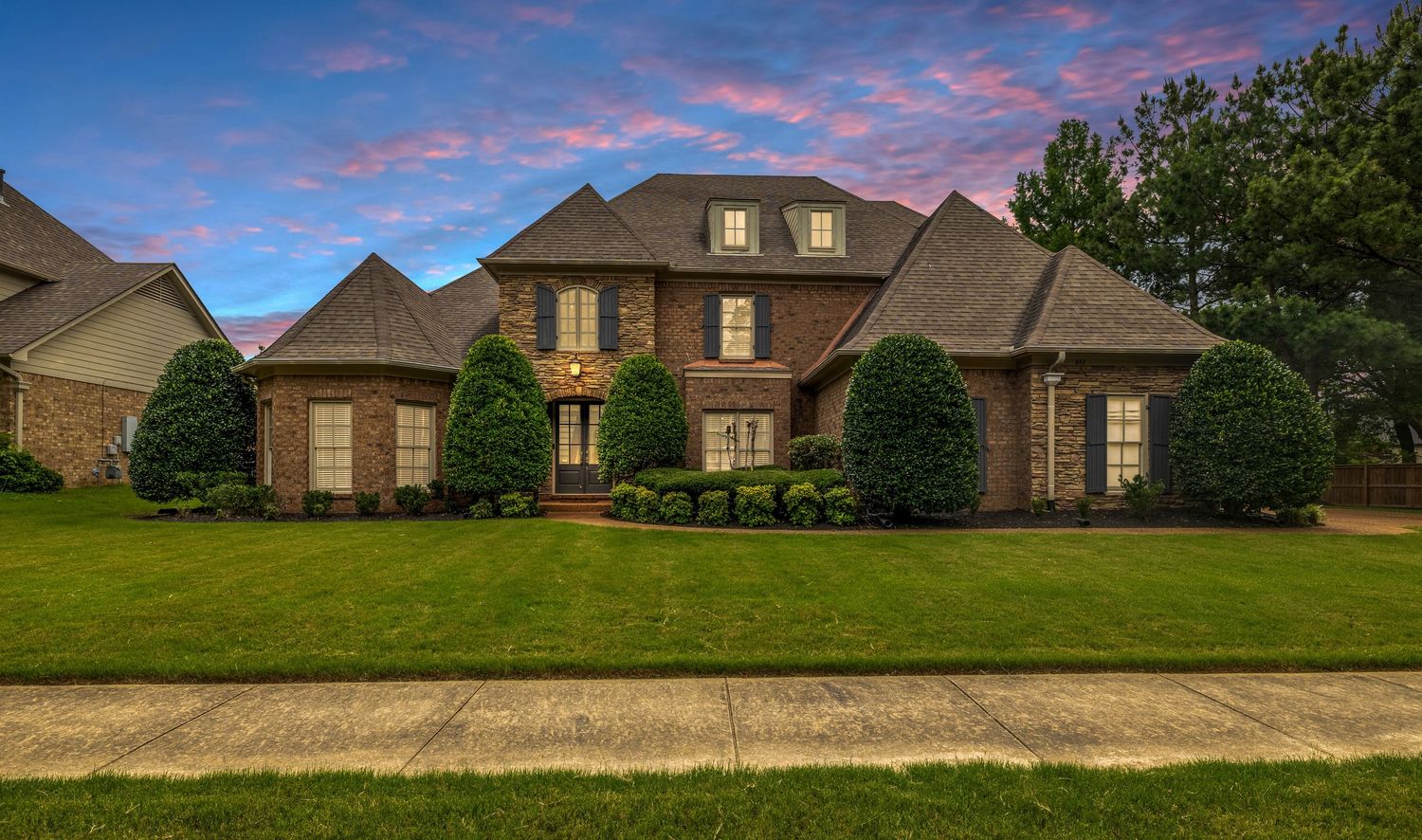 Stunning two-story brick home with lush green lawn and manicured shrubs at twilight with colorful pink sky.