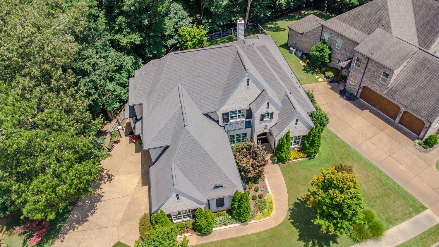 Aerial drone view of a modern gray house with multiple rooflines surrounded by landscaping and trees.