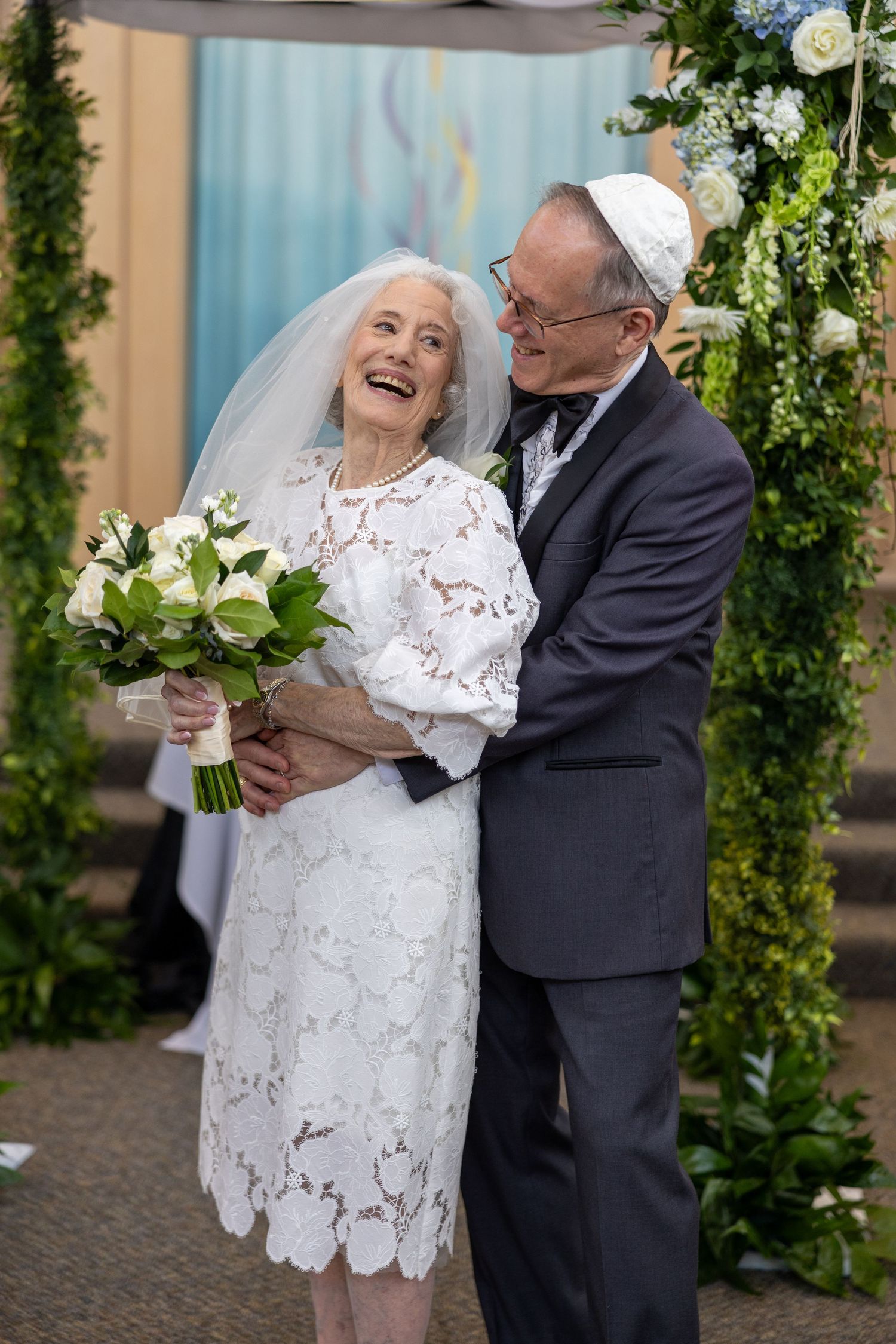 An elderly couple in wedding attire share joyful moments together during a photo session with floral decor.