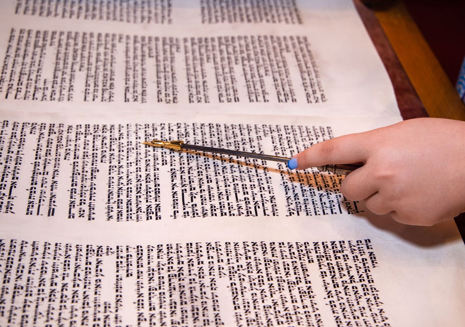 A hand holding a pencil points to text in a Hebrew religious manuscript or Torah scroll during study.