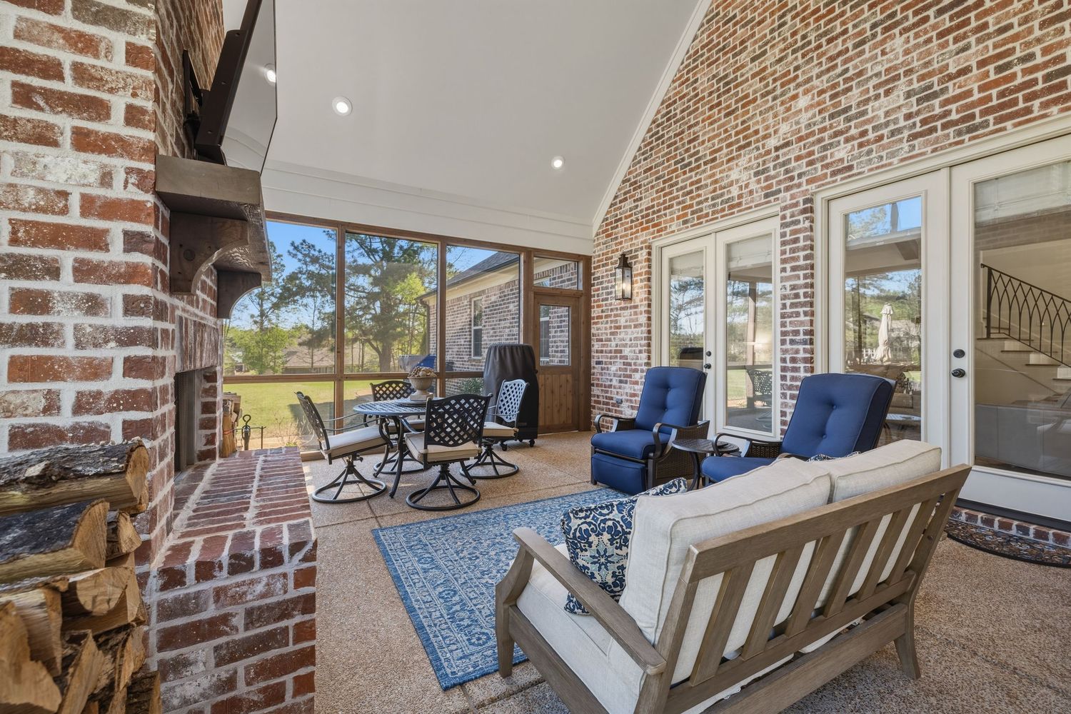 Covered brick patio with outdoor seating, blue accent chairs, fireplace, and French doors leading to a lush backyard.