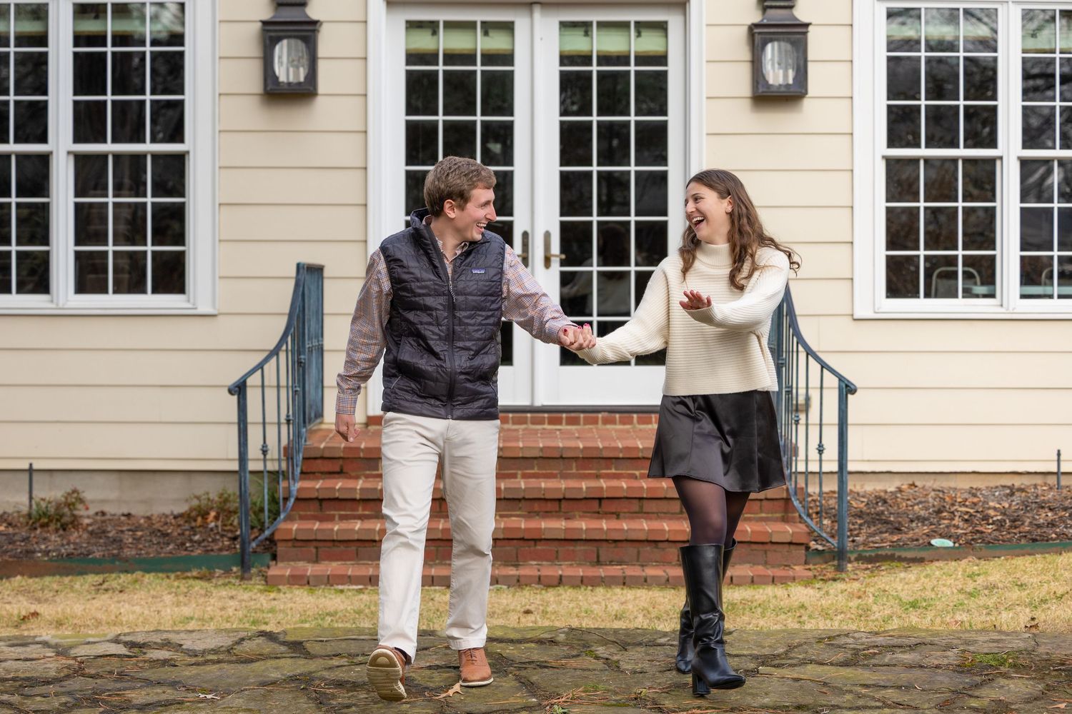 A couple shares a playful moment while standing on brick steps outside a traditional home with large windows.