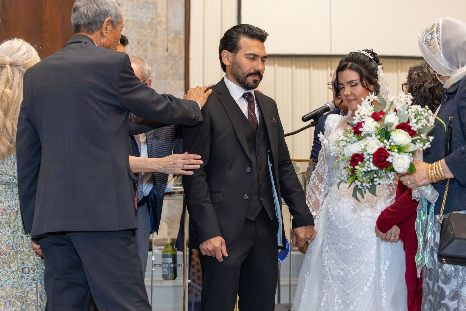 Wedding party gathers outside church entrance with bride in lace gown and red floral bouquet.