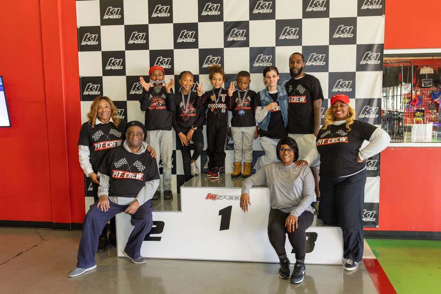 Group celebrating on a podium with checkered racing backdrop at an indoor event.