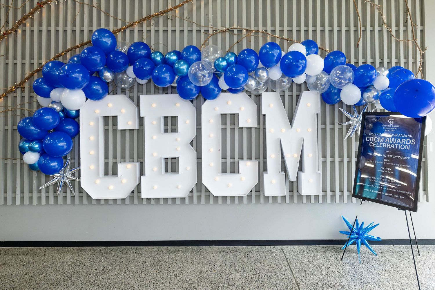 Large white CBCM letters mounted on wall with blue and white balloon arch decoration and promotional material display.