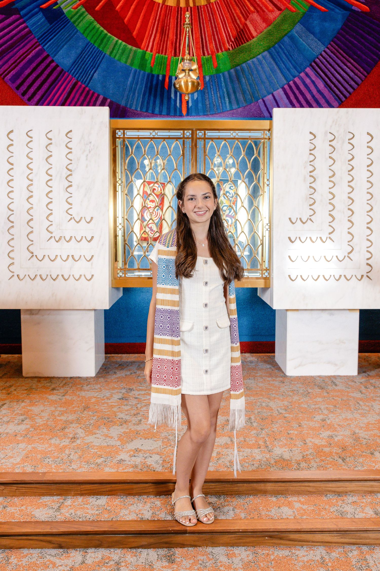 Young girl in white dress and colorful tallit stands smiling at her Bat Mitzvah ceremony in a vibrant synagogue.