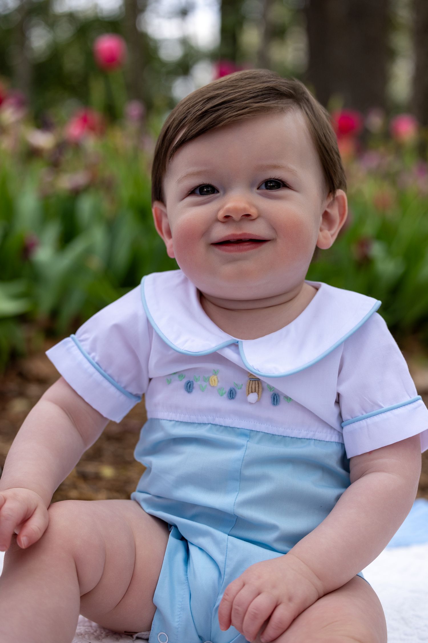 A baby in a white and blue outfit sits outdoors with pink flowers blooming in the background.