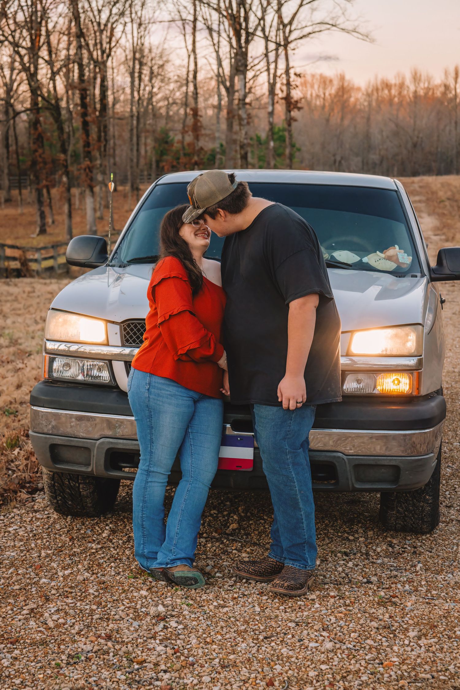Couple in casual attire leaning against a parked car at sunset with autumn trees in the background.
