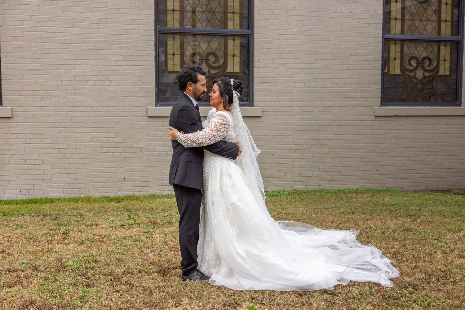 Wedding couple poses outside church with elegant stained glass windows and brick facade.