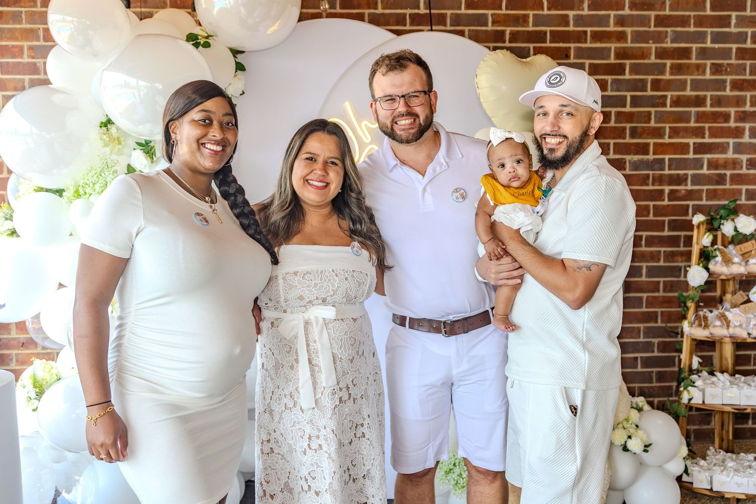 Group photo with white outfits and balloons against a brick wall background.