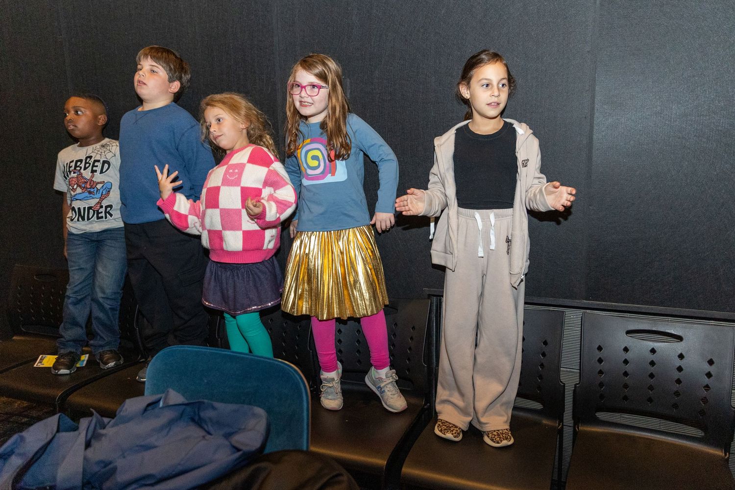 Children in casual clothes stand against a dark wall during a performance.