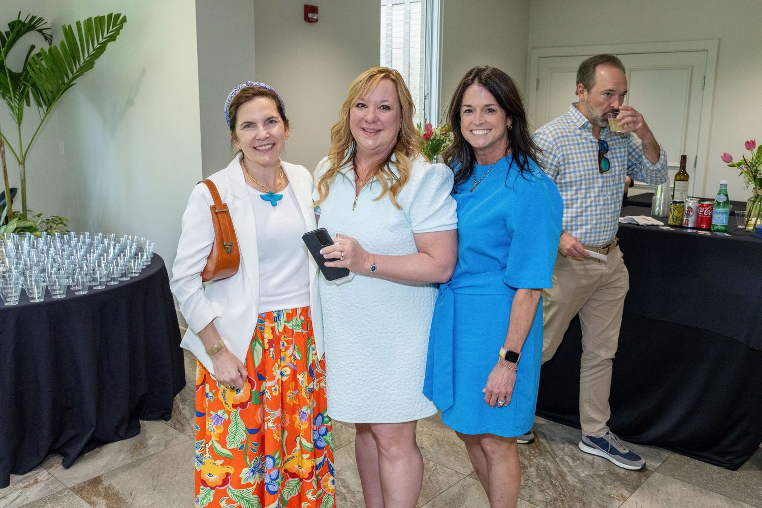 Three women smiling together at an indoor event with a bar and glassware display in the background.