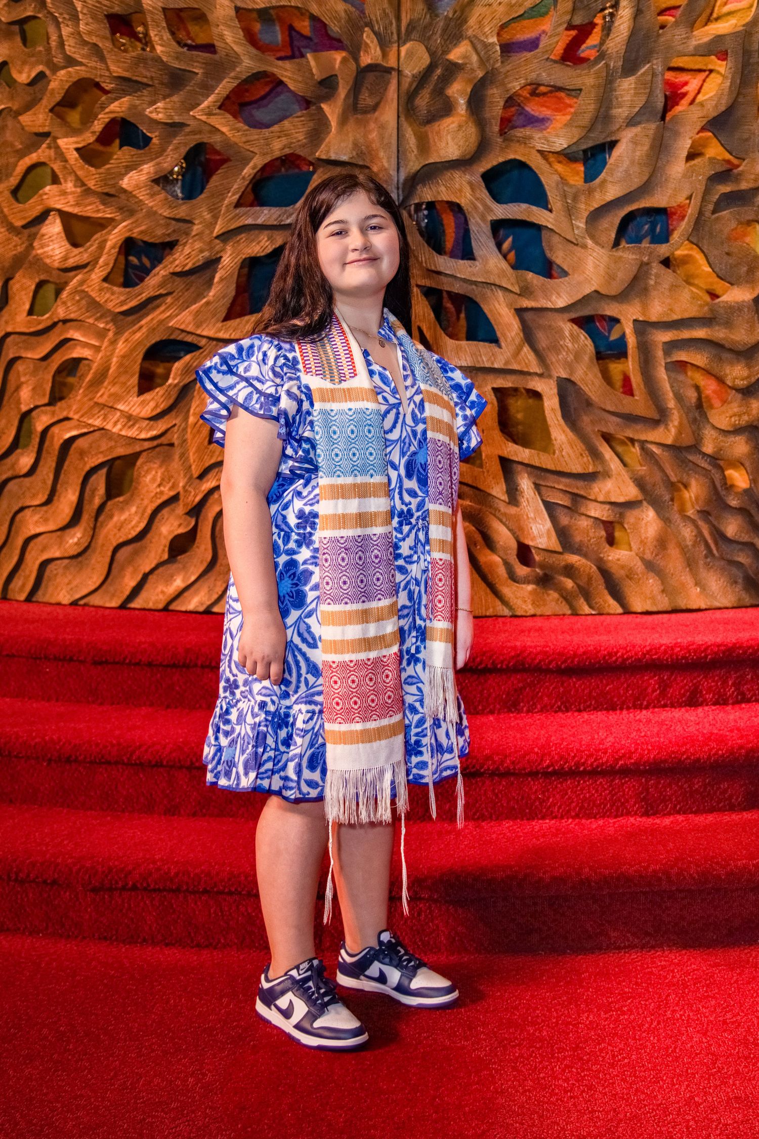 Child wearing blue and white patterned outfit poses on red carpet steps against decorative wooden wall background.