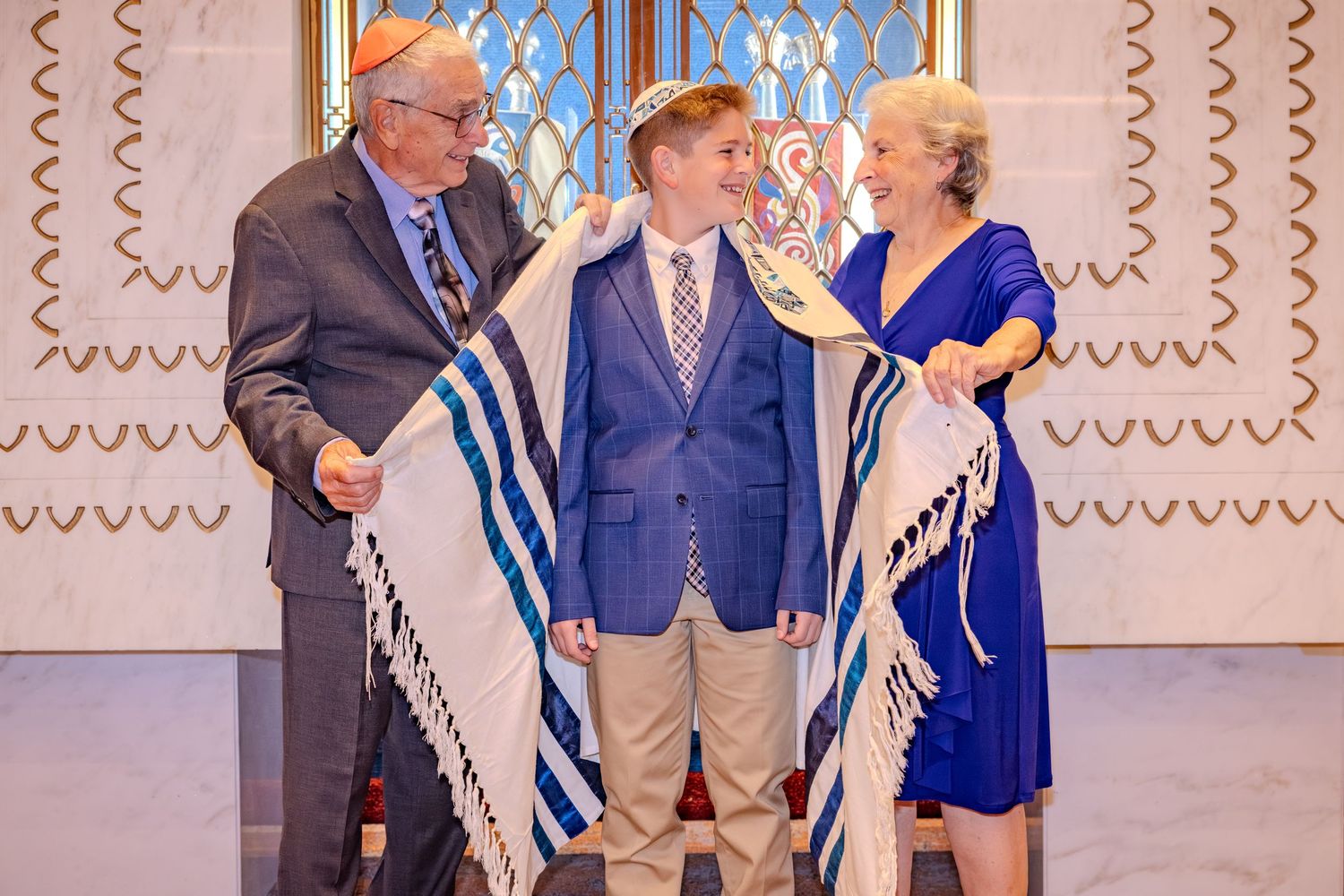 Boy in blue blazer at Bar Mitzvah ceremony, flanked by grandparents holding a tallit in a synagogue.