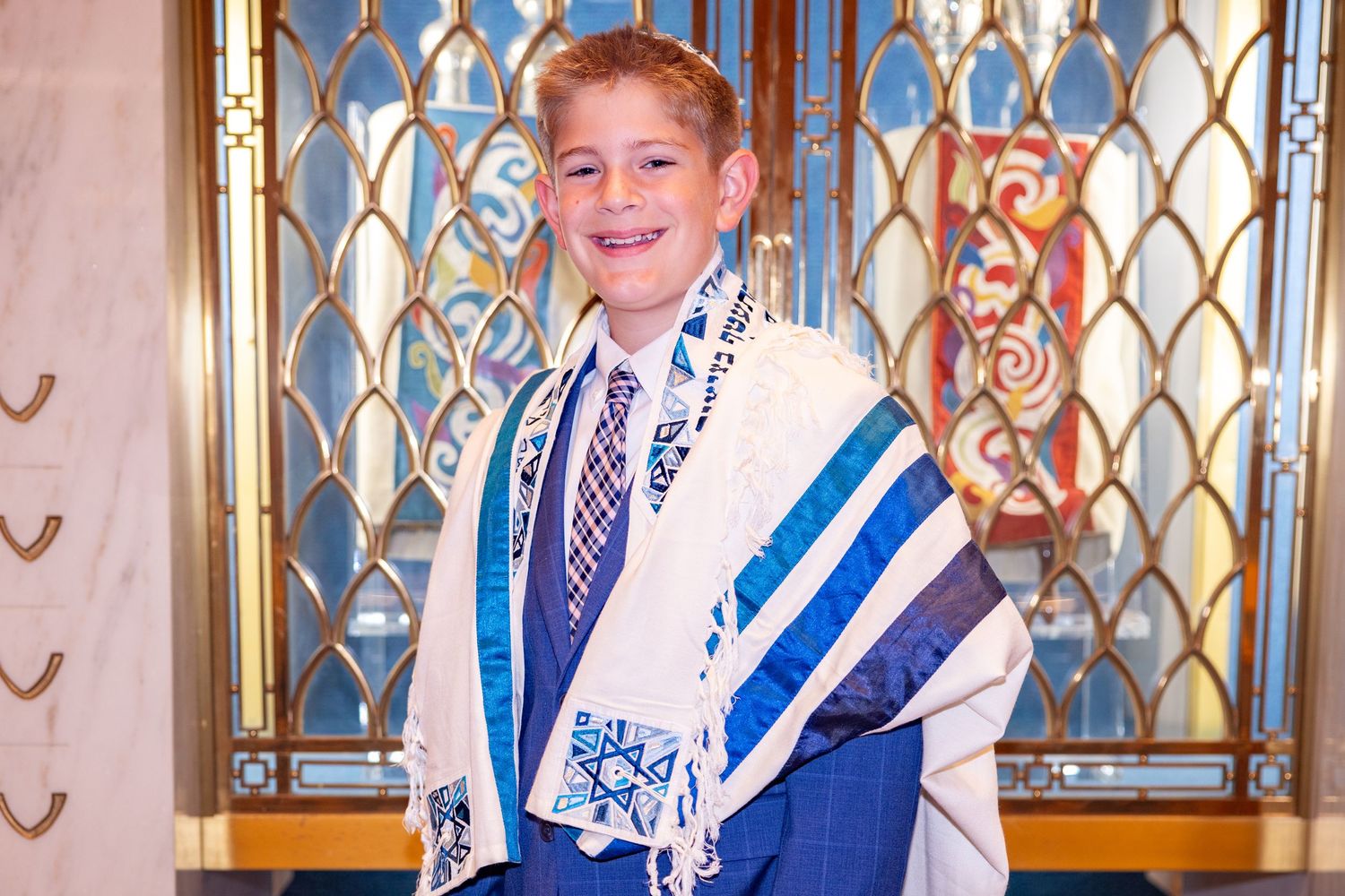 Young boy in bar mitzvah attire wearing a tallit with Star of David in a synagogue.