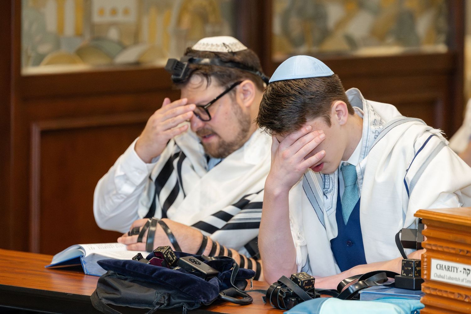 Two Jewish men in tallitot and kippot pray with heads bowed during a religious service in a synagogue.