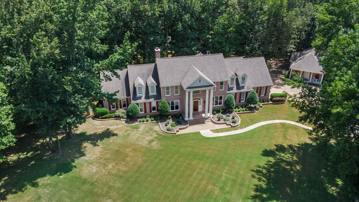Aerial drone panorama of residential neighborhood with large houses and green lawns surrounded by mature trees.
