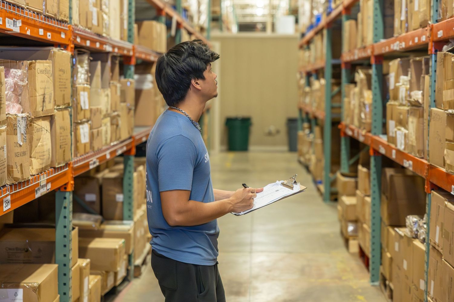 Workers checking inventory in warehouse aisle with orange metal shelving units.