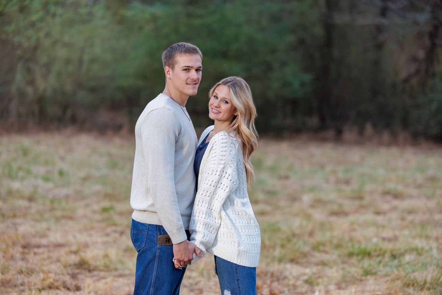 A couple in casual attire embraces while standing in a grassy outdoor field during an autumn photo session.