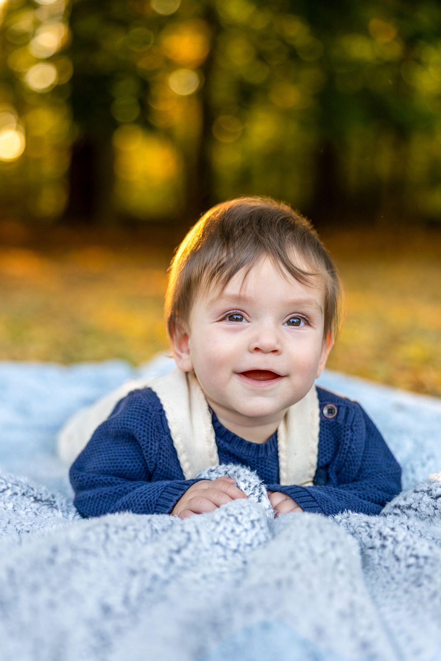 Baby lying on blanket outdoors with autumn foliage in background.
