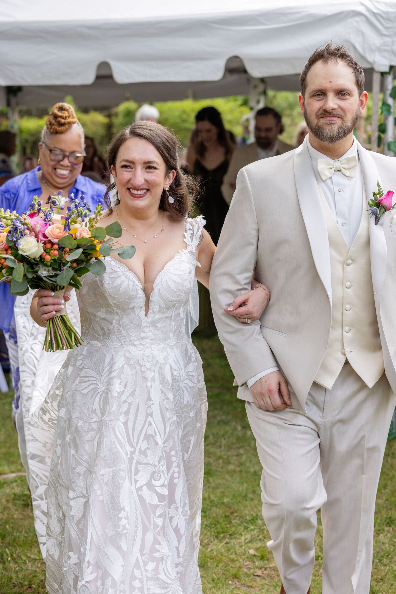 Smiling bride in lace gown holding colorful bouquet walks with groom in tan suit after outdoor wedding ceremony.