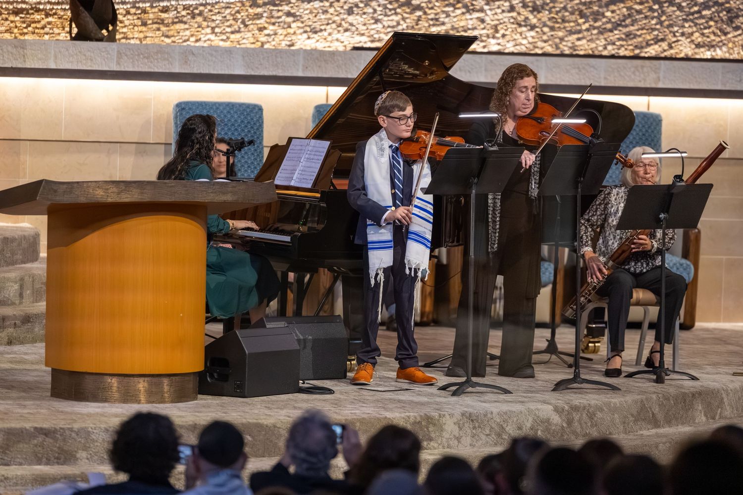 Musicians perform with violins and piano at a synagogue concert, with a young boy wearing a tallit.
