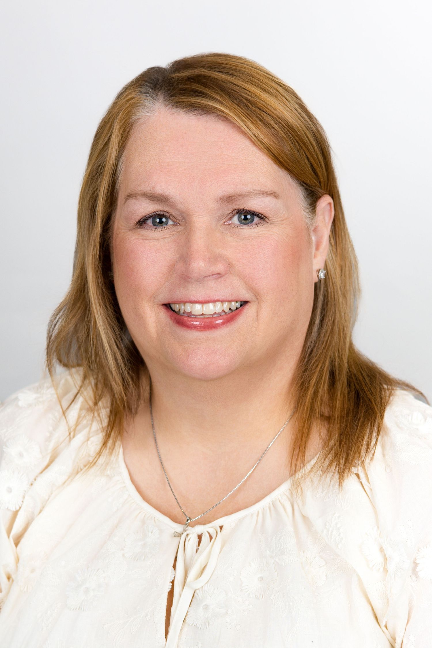 Professional headshot of someone wearing a white blouse and necklace against a light background, smiling warmly at camera.