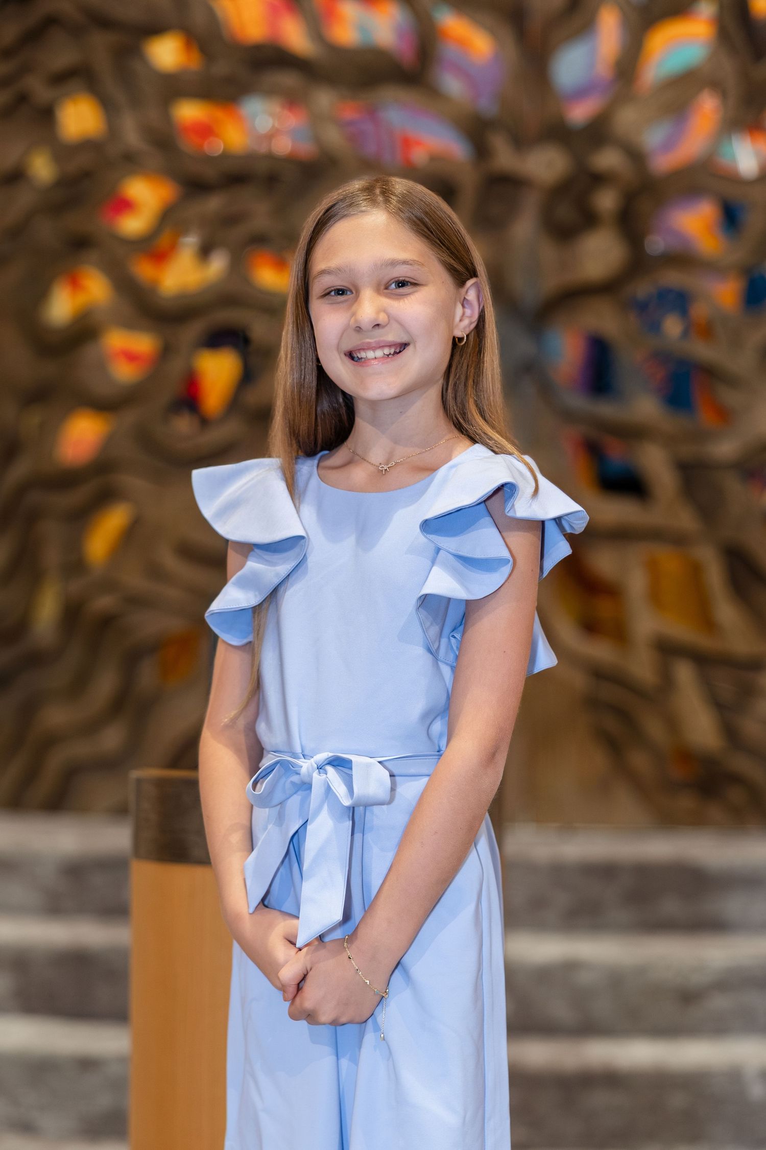 Smiling young girl in a light blue ruffle jumpsuit posing in a decorated religious venue with stained glass.