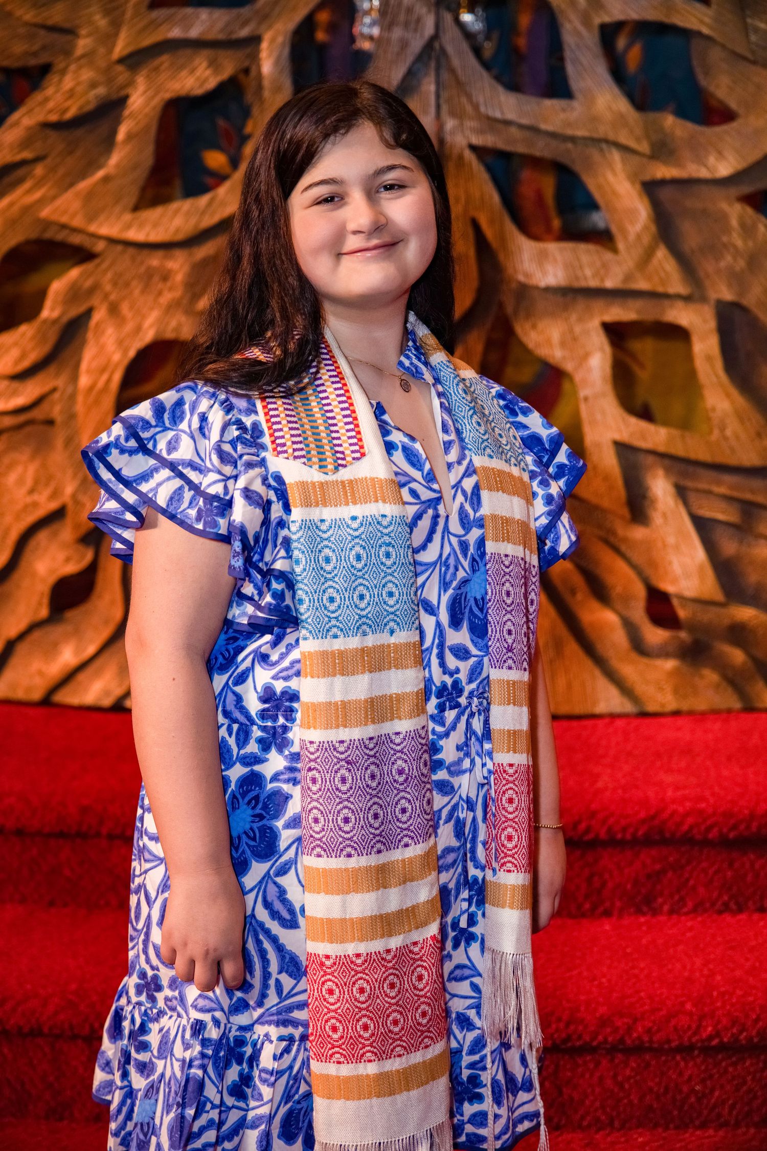 A young person in a blue and white floral dress poses against a backdrop of autumn leaves and red carpet.