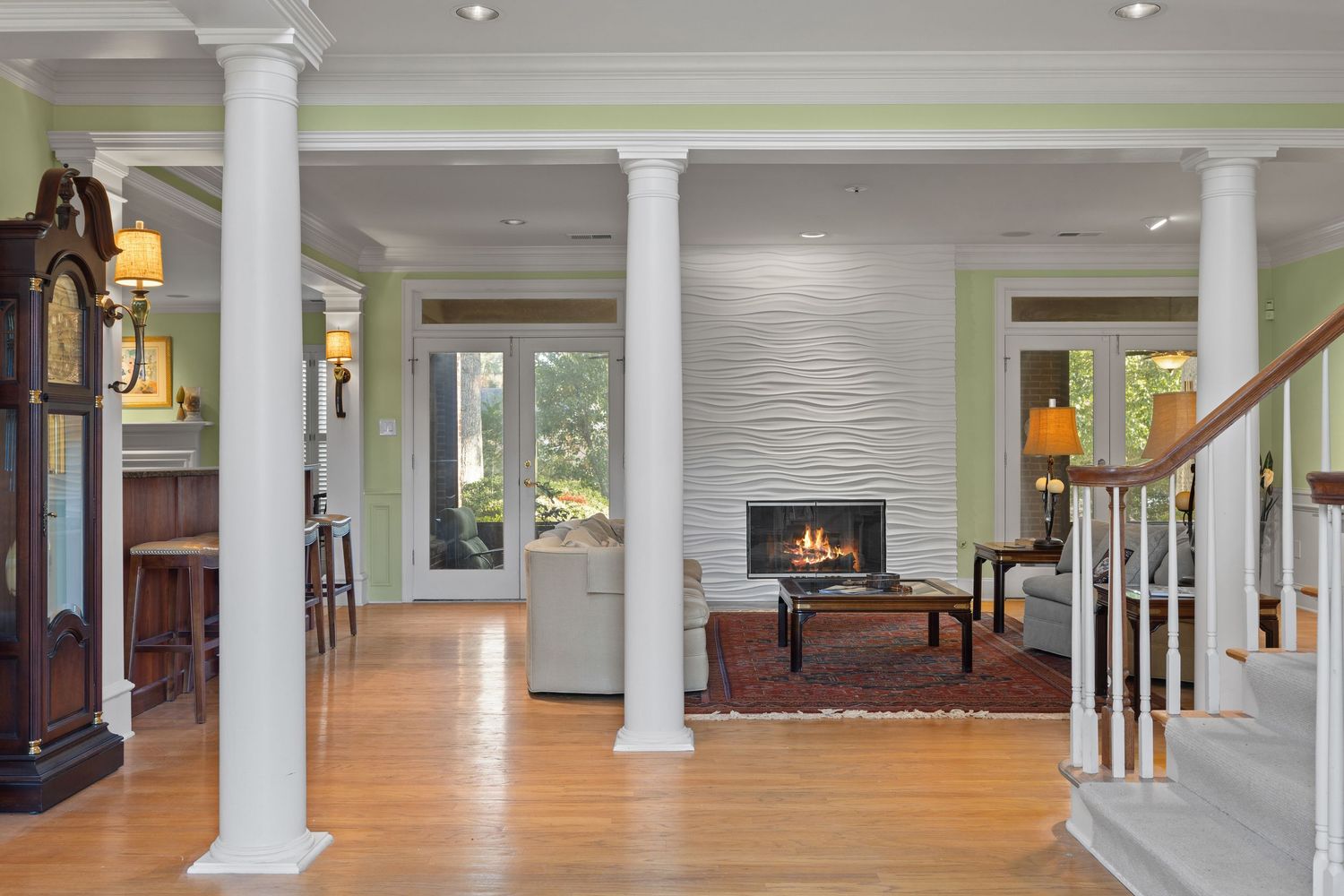 Modern open concept living space with white columns, hardwood floors, and a stone fireplace visible in the distance.