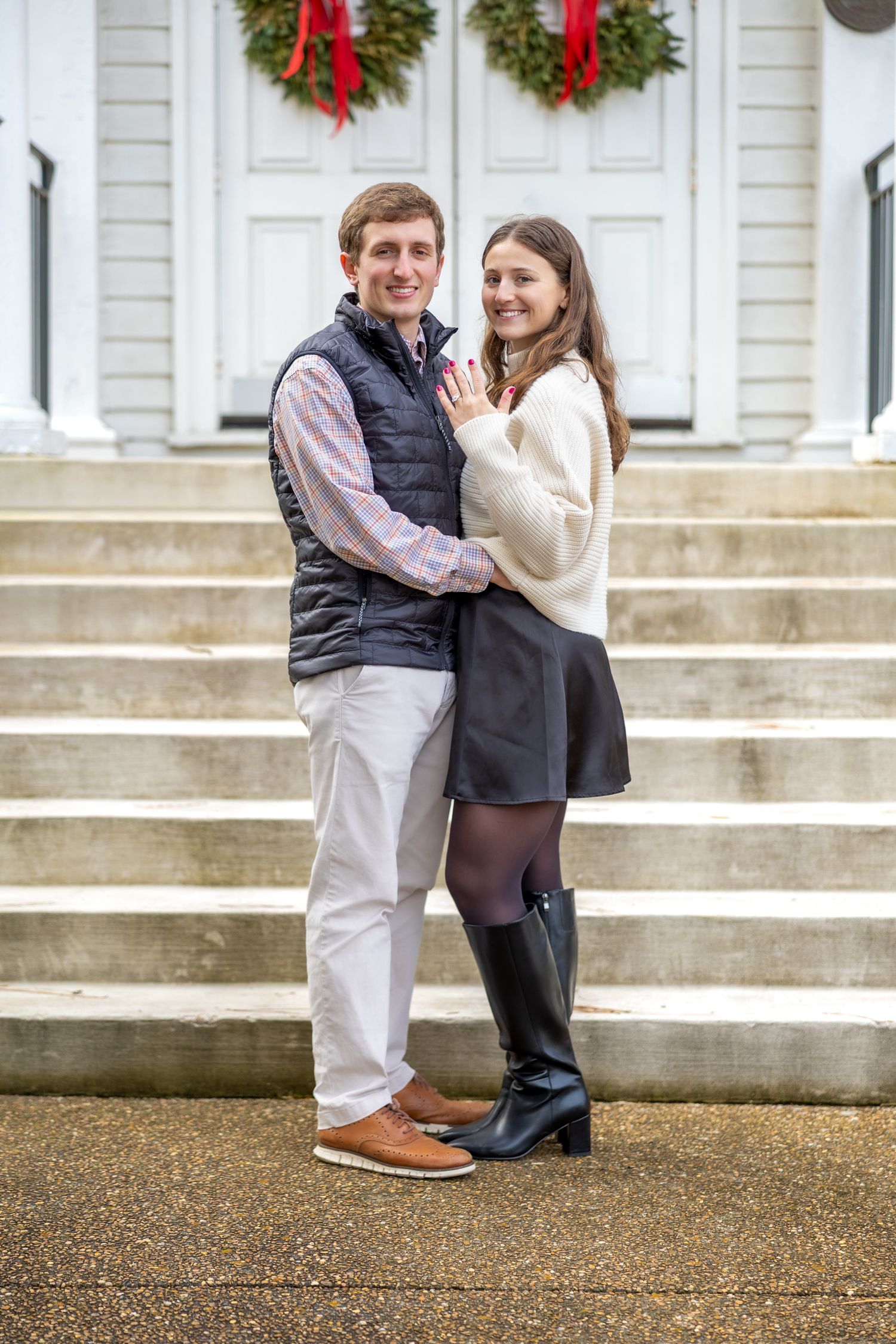 A couple embraces on white steps decorated with holiday wreaths during a winter photo session.