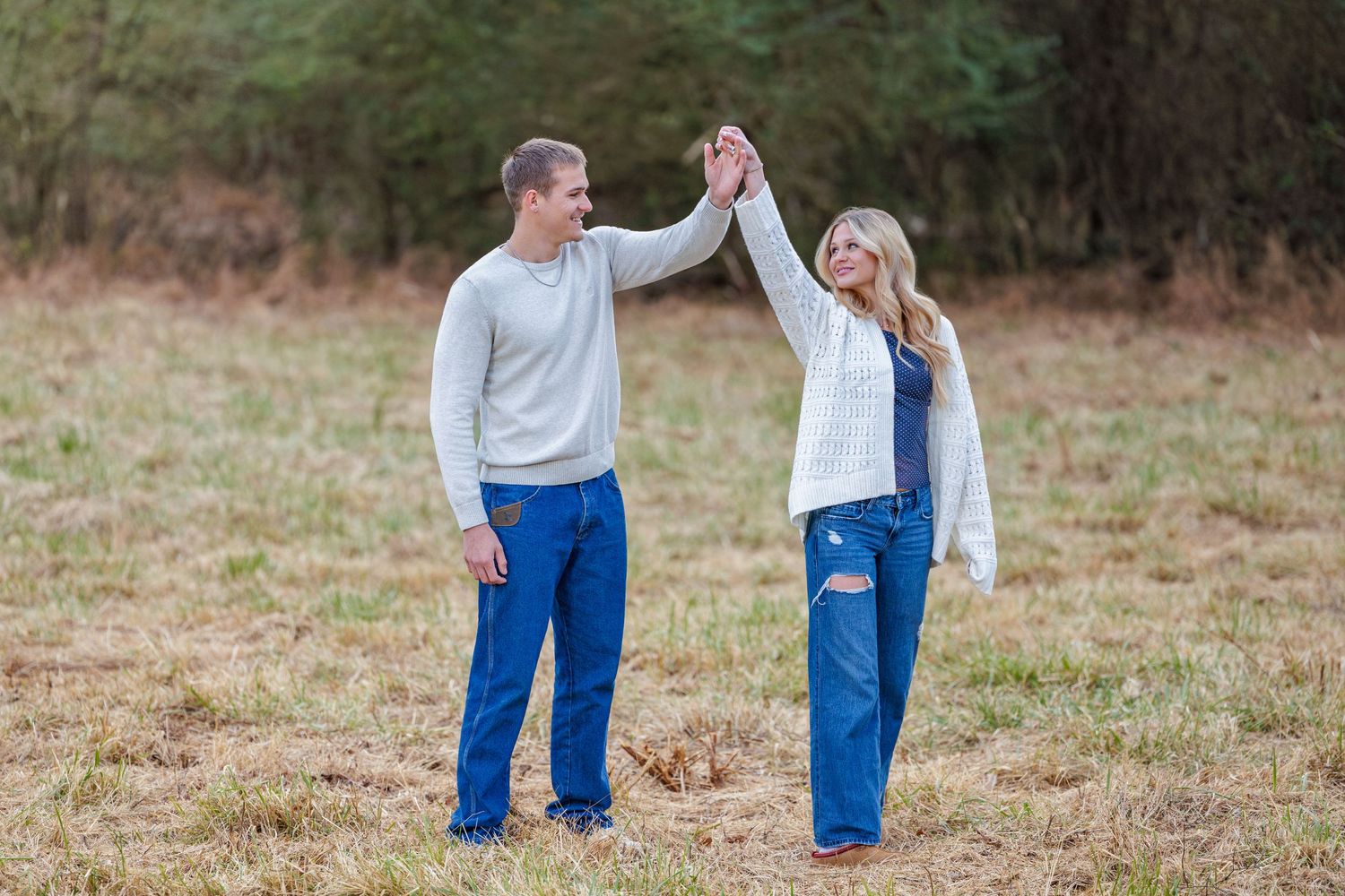 Two people in blue jeans and white tops hold hands while walking together in a grassy outdoor field.