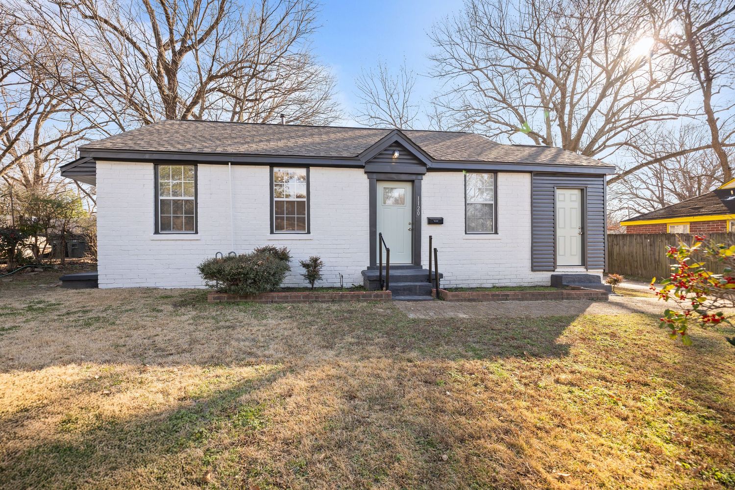 A panoramic view of a single-story ranch house with white siding and dark roof set against bare winter trees.