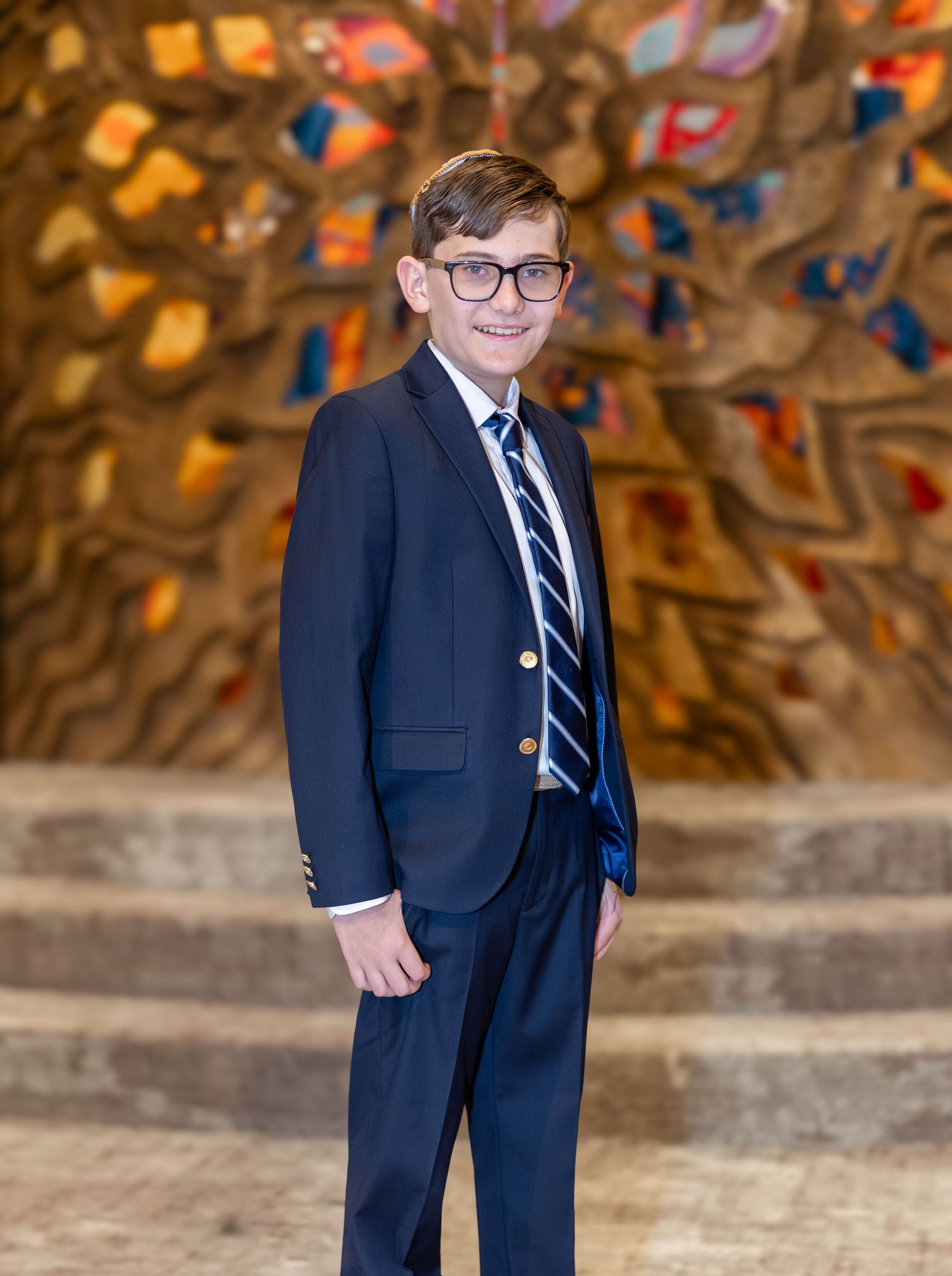 Young boy in navy blue suit and striped tie smiling in front of colorful mosaic wall backdrop.