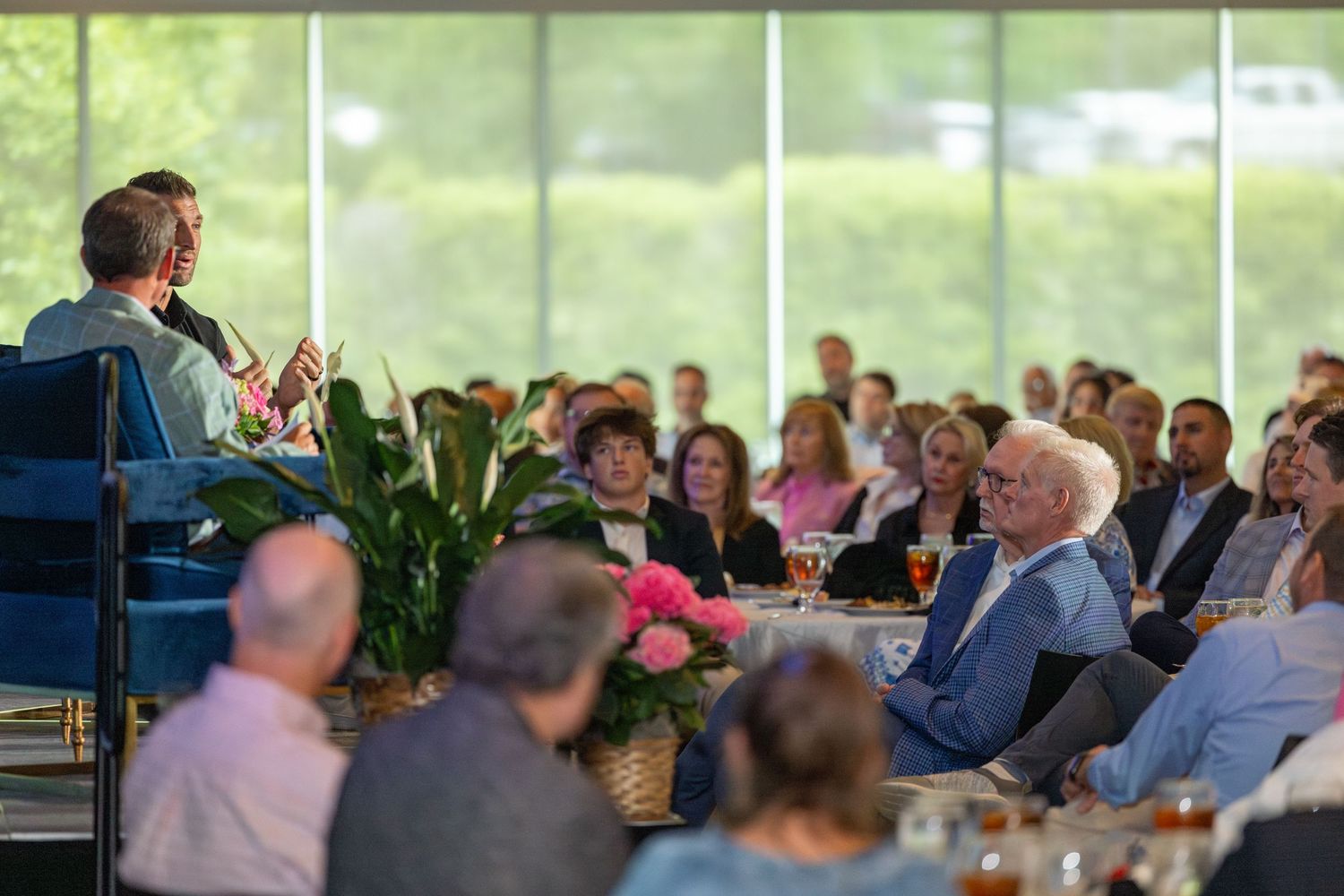 Speaker addresses a large audience at an elegant indoor event with floor-to-ceiling windows and floral centerpieces.