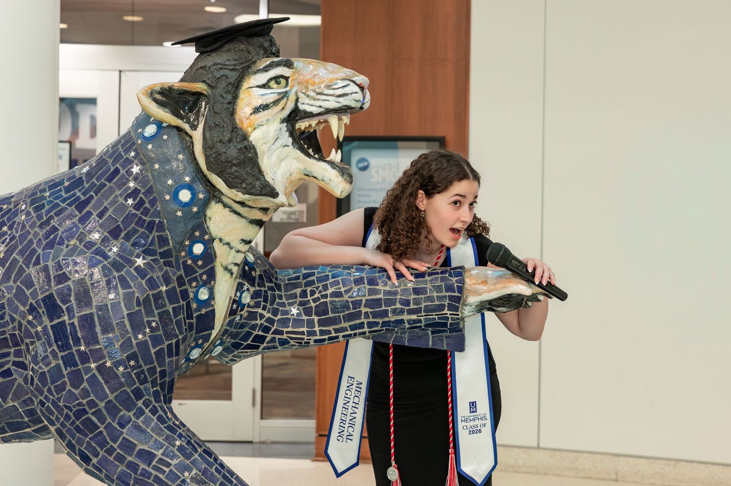 Graduate poses with mosaic tiger statue, wearing cap and gown with blue sash, holding microphone at university event.