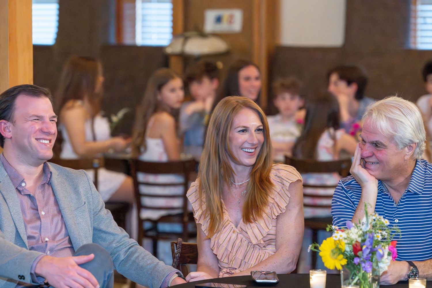 Three adults laughing and smiling together at a restaurant table with floral centerpiece and guests in background.