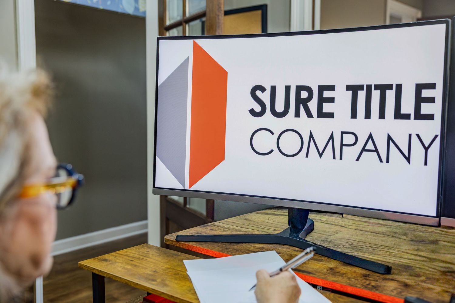 Someone writes notes at a wooden desk with the Sure Title Company logo displayed on a monitor screen.