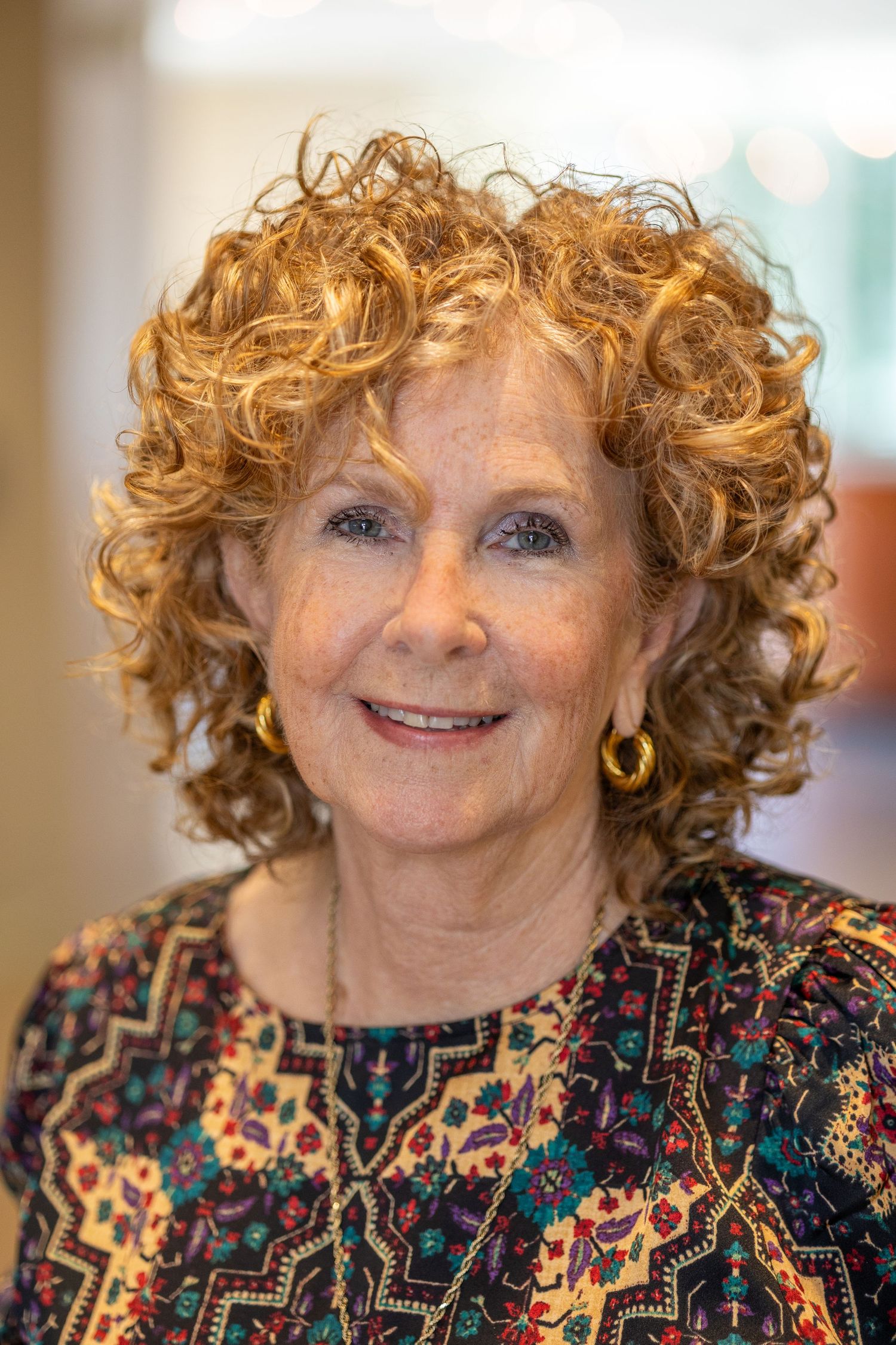 Close up portrait of woman with curly ginger hair wearing patterned top.